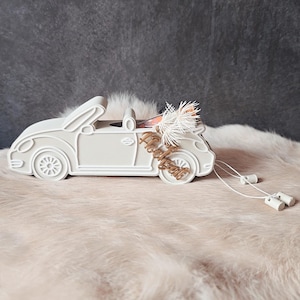 Könnte beinhalten: Ein weißes, cabrio-förmiges Dekorationsstück mit dem goldenen Schriftzug "Happily Ever After" an der Seite. Das Auto hat einen dekorativen Blumenschmuck hinten und zwei weiße Kordeln mit kleinen weißen Zylindern.