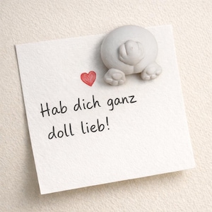 Könnte beinhalten: Ein weißer Haftzettel mit einem roten Herz und dem deutschen Text "Hab dich ganz doll lieb!". Ein grauer, runder, tierförmiger Magnet ist oben auf dem Zettel befestigt. Der Zettel liegt auf einer strukturierten, cremefarbenen Oberfläche.