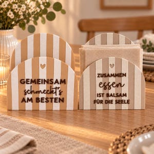 Op de afbeelding: Twee boogvormige servethouders met verticale beige en witte strepen. De linkerhouder toont de tekst GEMEINSAM schmeckt's AM BESTEN met een kleine hartvormige uitsparing. De rechterhouder toont ZUSAMMEN essen IST BALSAM FÜR DIE SEELE.