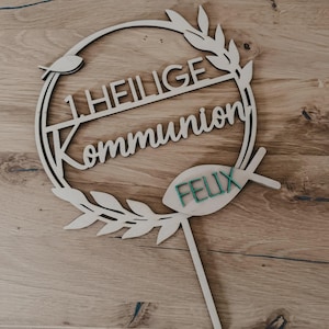 以下が含まれることがあります： 「1. HEILIGE Kommunion」の文字が円形にデザインされた木製のケーキトッパー。魚と「FELIX」という名前が緑色で描かれています。明るい色の木材で作られており、挿入用の棒が付いています。