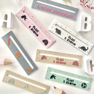 Puede incluir: Una colección de coloridas decoraciones rectangulares con temática escolar con centros recortados. Las decoraciones presentan el texto "ENDLICH SCHULKIND!" y "Du bist 1. Klasse", junto con ABC, arcoíris e iconos relacionados con la escuela.