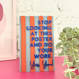 Könnte beinhalten: Ein rechteckiges Schild mit hellgrauem Hintergrund und vier vertikalen orangefarbenen Streifen. Der blaue Text lautet: STOP LOOKING AT THIS POSTER AND DO YOUR WORK. Yes, You! Das Schild steht aufrecht auf einer leuchtend pinken Oberfläche.