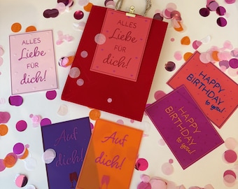 Può includere: Un sacchetto regalo rosso con manico dorato e un biglietto con la scritta "Alles Liebe für dich!" è circondato da biglietti con la scritta "Happy Birthday to you!" e "Auf dich!" con coriandoli.