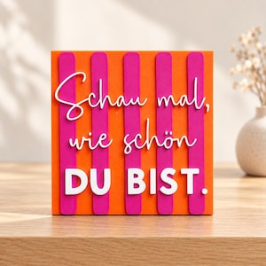 Könnte beinhalten: Ein quadratisches Dekoschild mit vertikalen orangefarbenen und pinken Streifen. Der weiße Schriftzug lautet "Schau mal, wie schön DU BIST.". Das Schild hat eine geschichtete, dreidimensionale Optik mit erhabenen weißen Buchstaben auf dem gestreiften Hintergrund.