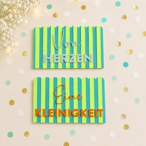 Puede incluir: Dos letreros rectangulares con borde superior festoneado y rayas verticales verde lima y azul cerceta. El letrero superior muestra el texto Von en letra cursiva azul claro y HERZEN en letras de bloque azul claro. El letrero inferior muestra Eine en letra cursiva naranja y KLEINIGKEIT en letras de bloque naranja.