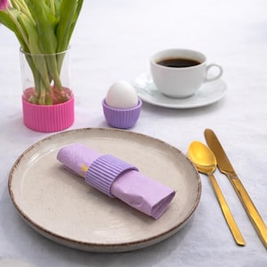 Może przedstawiać: Nakrycie stołu śniadaniowego z beżowym talerzem, złożoną lawendową serwetką z lawendowym pierścieniem na serwetki i złotymi sztućcami. Biała filiżanka kawy, fioletowy kubek na jajko z jajkiem i wazon z tulipanami dopełniają scenę.