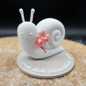 Pode incluir: Uma figura de caracol branco texturizado com um rosto sorridente e um buquê de flores cor-de-rosa. O caracol está sobre uma base circular cinza com texto branco. Um item decorativo caprichoso.