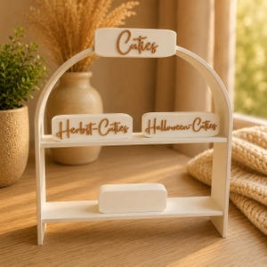 Könnte beinhalten: Ein weißes, zweistöckiges, bogenförmiges Regal mit drei rechteckigen Schildern mit brauner Schrift. Das obere Schild trägt die Aufschrift Cuties, das mittlere linke Herbst-Cuties und das mittlere rechte Halloween-Cuties. Ein leerer weißer Block steht auf der unteren Ablage.