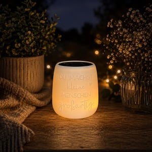 May include: A glowing white solar-powered lantern with the German text "Du machst kleine Menschen großartig" is displayed on a wooden surface. The lantern is cylindrical with a slightly flared top. A potted plant and a vase of flowers are in the background.