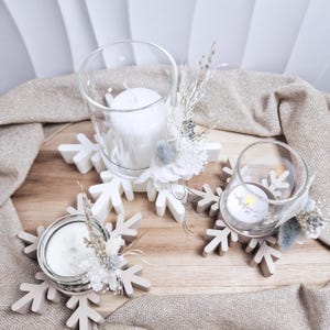 Pode incluir: Três castiçais com velas brancas e arranjos florais decorativos. As velas estão em suportes de vidro transparente, colocados em bases em forma de floco de neve. As bases são de cor bege claro, o tema é decoração de inverno ou feriado.