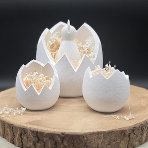 Könnte beinhalten: Drei weiße, eierschalenförmige Keramikbehälter mit gezackten Kanten stehen auf einer Holzoberfläche. Das größte Ei enthält eine weiße Kükenfigur und Trockenblumen. Die anderen beiden Eier enthalten ebenfalls Trockenblumen.