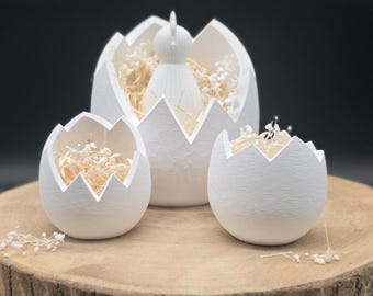 May include: Three white ceramic eggshell-shaped containers, each with a jagged edge, sit on a wooden surface. The largest egg holds a white chick figurine and dried flowers. The other two eggs also contain dried flowers.