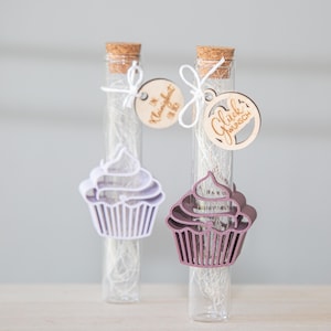 以下が含まれることがあります： コルク栓付きのガラス管2本。それぞれ白いフィラーとカップケーキ型の切り抜きが入っています。切り抜きはラベンダー色とモーブ色です。木製のタグにテキストが書かれており、白い紐でチューブに結ばれています。タグには「Glückwunsch」と「Kleinigkeit」と書かれています。