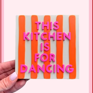 Könnte beinhalten: Ein quadratisches Wandschild mit hellgrauem Hintergrund und vertikalen orangefarbenen Streifen. Der erhabene rosa Schriftzug lautet THIS KITCHEN IS FOR DANCING, wobei die Wörter in fünf Zeilen in der Mitte der gestreiften Fläche angeordnet sind.