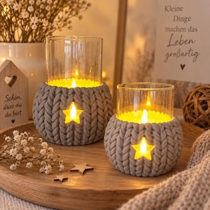 Può includere: Due portacandele in vetro rivestiti con coperture in maglia grossa grigio chiaro, ciascuno con un'apertura a forma di stella. All'interno, una calda luce LED illumina lo spazio. I portacandele poggiano su un vassoio in legno con stelle.