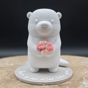 May include: A white otter figurine holding a small bouquet of pink flowers. The otter is standing on a gray circular base with the text "There is no otter like you" in a cursive font. The figurine has a textured surface.