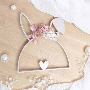 Könnte beinhalten: Eine weiße Hasen-Wanddekoration mit herzförmiger Nase. Der Hase ist mit einem rosa Band und einem Cluster aus Trockenblumen in Rosa- und Weißtönen verziert. Die Dekoration befindet sich vor einem hellen Holzhintergrund.