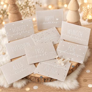 May include: A collection of rectangular light beige cards featuring white embossed text and heart or dot patterns. The cards display various German phrases including Happy Birthday, Wunscherfüller, Konfettiii für dich, Gutschein, Freu post, Für dich, Alles Liebe, and Post für dich.