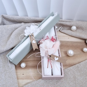 Może przedstawiać: Dwa prostokątne pudełka, jedno jasnozielone i jedno różowe, każde zawierające białe świece. Pudełka ozdobione są białymi kwiatami, różowymi akcentami i małymi etykietami w kształcie serca. Rozrzucone perły.