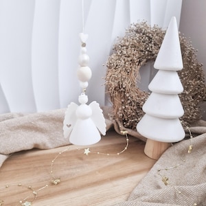 Może przedstawiać: Biała ozdoba w kształcie anioła z wycięciami w kształcie serca wisi na sznurku z koralików i zawieszki w kształcie gwiazdy. Widoczne są również biała, warstwowa choinka i suchy wieniec, sugerujące świąteczny motyw.