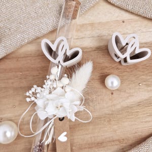 Peut inclure: Un flacon en verre transparent avec un bouchon en liège, orné de fleurs séchées blanches et d'une petite étiquette. Deux emporte-pièces en forme de cœur et deux perles sont disposés sur une surface en bois.