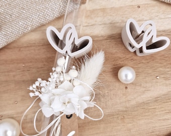 Pode incluir: Um tubo de ensaio de vidro transparente com rolha de cortiça, adornado com flores secas brancas, uma pequena etiqueta de papel castanho com um ícone de coração branco e um ornamento de coração duplo. Dois corações e duas pérolas brancas.