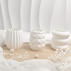 Op de afbeelding: Drie witte decoratieve vazen met verschillende geometrische ontwerpen. De vaas links heeft verticale lijnen, de middelste vaas heeft een ruitpatroon en de vaas rechts heeft een ontwerp van gestapelde ringen. De vazen staan op een houten oppervlak.