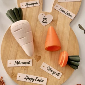 Op de afbeelding: Een houten paashaasvormig bord met een lichtroze wortel, een oranje wortel en een wortelschijf. Op het bord staan de zinnen "Happy Easter", "Hasenpost", "Mohrenpost", "Frohe Ostern", "Ostergruß" en "Für dich".