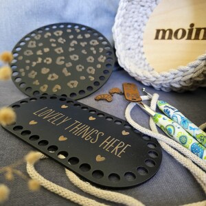 Könnte beinhalten: Eine schwarze ovale Basis mit dem Text "LOVELY THINGS HERE" und goldenen Herzen sowie eine schwarze runde Basis mit Leopardenmuster. Häkelnadeln mit gemusterten Griffen und eine graue gehäkelte Tasche sind ebenfalls zu sehen.