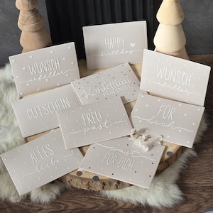 Könnte beinhalten: Eine Sammlung hellrosa Geschenkkarten mit weißem, geprägtem Text und Herzakzenten. Die Karten zeigen Sätze wie "Happy Birthday", "Für dich" und "Alles Liebe". Sie sind auf einer Holzoberfläche angeordnet.