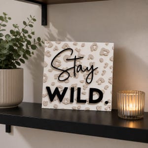 May include: A square white sign featuring a raised beige leopard print pattern. The word Stay is written in a black cursive script font, and the word WILD. is written below in a bold, black, sans-serif capital letter font.