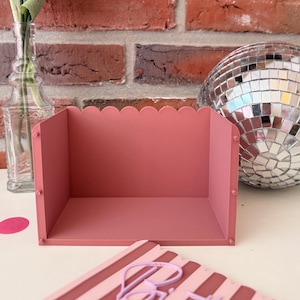 May include: A dusty rose, open-front storage bin with a scalloped top edge. The rectangular container features small, circular pegs on the front side panels and is constructed from a solid, matte-finish material with a flat base.