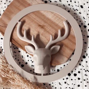 Könnte beinhalten: Eine matte beige Hirschkopfskulptur, die in einem kreisförmigen Rahmen montiert ist. Der Hirsch weist detaillierte Geweihe und Ohren auf und ist in einem soliden, neutralen Farbton gehalten. Der kreisförmige Rahmen verbindet sich oben und unten.