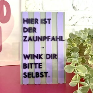 Könnte beinhalten: Ein rechteckiges Schild mit hellgrünem Hintergrund und vier vertikalen hellvioletten Streifen. Dunkelviolette erhabene Buchstaben bilden den deutschen Text: HIER IST DER ZAUNPFAHL - WINK DIR BITTE SELBST. Das Schild hat ein flaches Design.