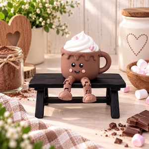 Könnte beinhalten: Eine braune Tasse mit lächelndem Gesicht, rosa Wangen und beweglichen Beinen, die auf einer kleinen schwarzen Bank sitzt. Die Tasse ist mit weißer Schlagsahne und rosa Marshmallow-Stückchen garniert und hat einen Henkel an der rechten Seite.