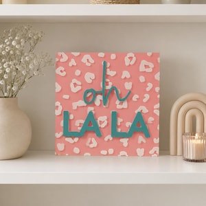 Puede incluir: Un letrero cuadrado de madera con fondo rosa coral y un patrón de estampado de leopardo blanco. El letrero muestra el texto en color verde azulado "oh LA LA" en una combinación de letra cursiva y de molde, con "oh" en cursiva sobre "LA LA".