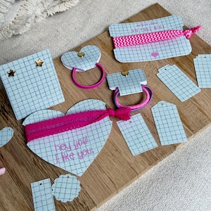 May include: A collection of hair accessories and tags on a wooden surface. The items include heart-shaped and rectangular cards with pink elastic hair ties, some with text. The cards have a grid pattern and are adorned with gold star embellishments.