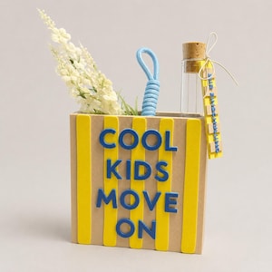 Op de afbeelding: Een beige rechthoekige houder met verticale gele strepen en de blauwe tekst COOL KIDS MOVE ON. Het bevat een lichtblauw touw als handvat, witte bloemstelen en een klein glazen flesje met een kurk en een geel label met de tekst EINZELSTÜCK.