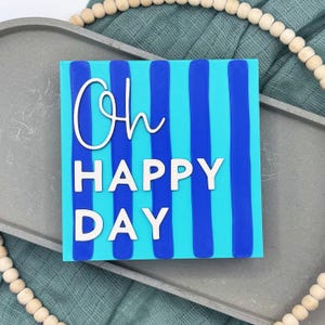 Peut inclure: Une plaque carrée présentant des rayures verticales alternant entre le bleu vif et le cyan. Le texte "Oh" est écrit en lettres cursives blanches, tandis que les mots "HAPPY DAY" apparaissent en dessous dans une police blanche sans empattement.