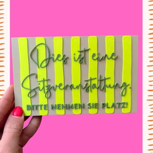 May include: A rectangular sign with a beige background and vertical neon yellow stripes. The sign features dark green text in a cursive script that reads "Dies ist eine Sitzveranstaltung." and block letters that read "BITTE NEHMEN SIE PLATZ!"