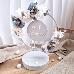 May include: A white metal floral wreath with a circular sign that reads "lich willkommen". The wreath is adorned with white, blue, and gray dried flowers and foliage. The base is white with a heart detail.