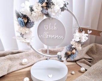 May include: A white metal floral wreath with a circular sign that reads "lich willkommen". The wreath is decorated with white, blue, and grey dried flowers and foliage. The base is white with a heart detail.