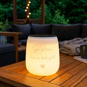 Puede incluir: Una linterna blanca brillante con el texto alemán "So schön, dass es dich gibt!" y un pequeño corazón rosa. La linterna está sobre una mesa de madera, con muebles de exterior y luces de cadena en el fondo.