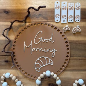 Könnte beinhalten: Ein braunes rundes Schild mit den Worten "Good Morning" und einem weißen Croissant-Design. Das Schild hat eine Reihe kleiner Löcher am Rand. Es gibt auch weiße und braune Croissant-förmige Ausschnitte und eine Perlenkette.