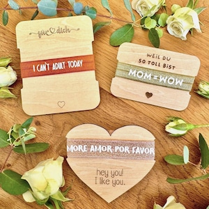 Könnte beinhalten: Geschenkanhänger aus Holz mit Aufschriften wie "I CAN'T ADULT TODAY" und "MOM=WOW". Die Anhänger haben dekorative Bänder und Herzformen. Die Anhänger liegen auf einer Holzoberfläche mit Blumen.