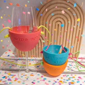 Könnte beinhalten: Ein klares Weinglas mit einem roten Silikonhalter mit der Aufschrift "wine not?" ist abgebildet. Zwei zusätzliche Silikonhalter, einer blau und einer orange, sind gestapelt. Der blaue Halter trägt die Aufschrift "partners in wine". Bunte Konfetti sind verstreut.