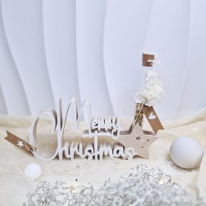 Puede incluir: Adorno navideño con las palabras "Merry Christmas" en blanco. Un vial de vidrio con flores blancas y un tapón de corcho se encuentra sobre una base en forma de estrella. Incluye una pequeña bola blanca y una etiqueta marrón.