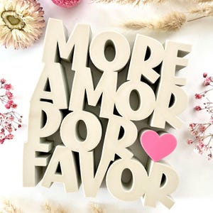 Könnte beinhalten: Ein cremefarbenes Dekoschild mit den Worten "MORE AMOR POR FAVOR" in gestapelten Blockbuchstaben. Ein kleines, rosa Herz befindet sich neben dem Wort "AMOR". Das Schild ist von Trockenblumen und Grün umgeben.