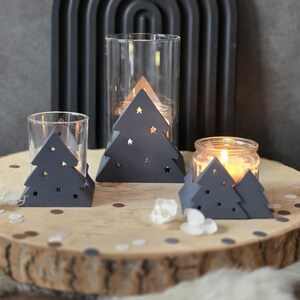 May include: A festive arrangement of candle holders. The image shows gray tree-shaped candle holders with star cutouts, glass votives, and a jar candle, displayed on a wooden slice. A dark gray arch is in the background, creating a seasonal atmosphere.