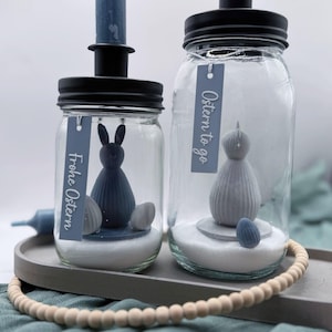 Könnte beinhalten: Zwei Gläser mit schwarzen Deckeln, jeweils mit einer Osterdekoration. Ein Glas zeigt einen blauen Hasen und Eier, das andere eine graue Figur und ein Ei. Beide haben Etiketten mit "Frohe Ostern" und "Ostern to go".